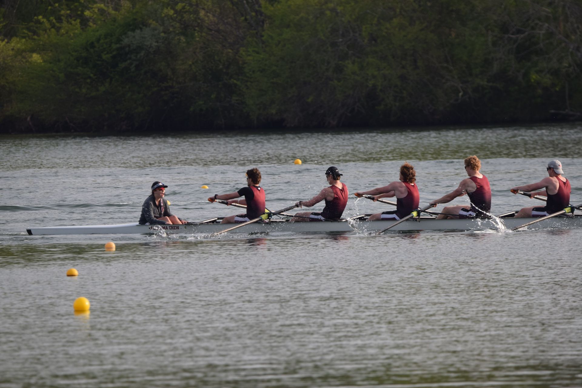 A rowing team in maroon uniforms races on a river. A coxswain steers, while the rowers propel the boat forward.