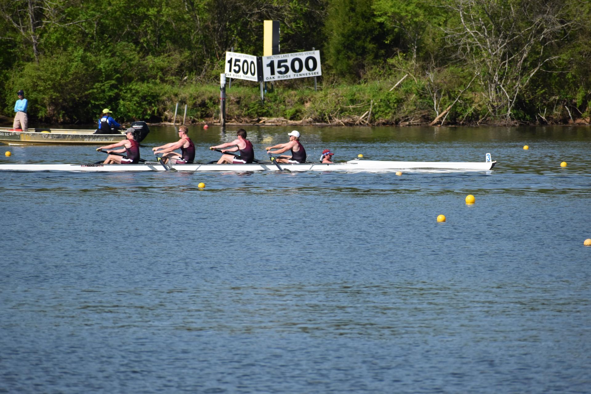 Rowing team racing on a river, approaching the 1500-meter mark, with trees in the background.