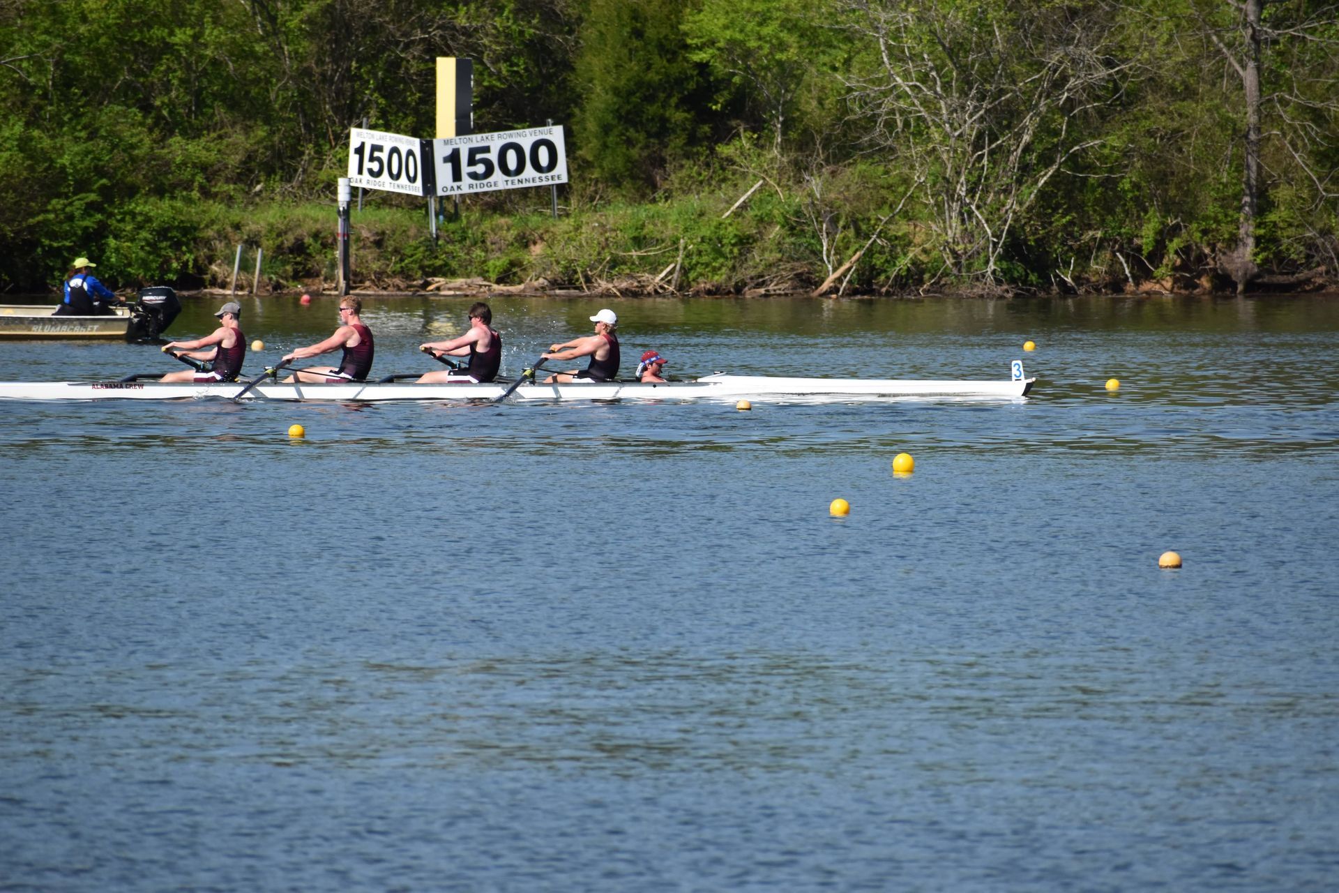 Three rowers in a boat on a river race towards the finish line, with a 