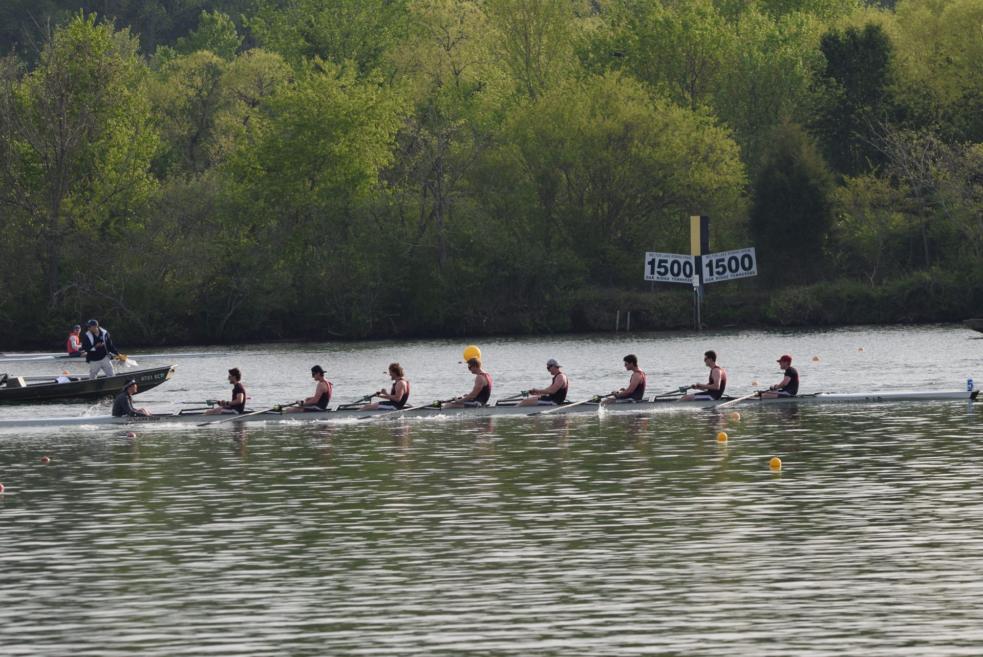 Rowing team in a race on a river; trees in the background; a support boat is beside them.