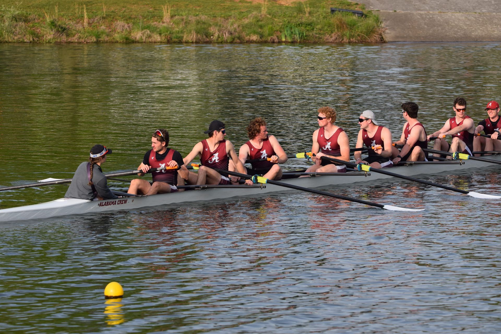 Rowing team in a boat. The coxswain faces forward, others row, and a yellow buoy is visible.