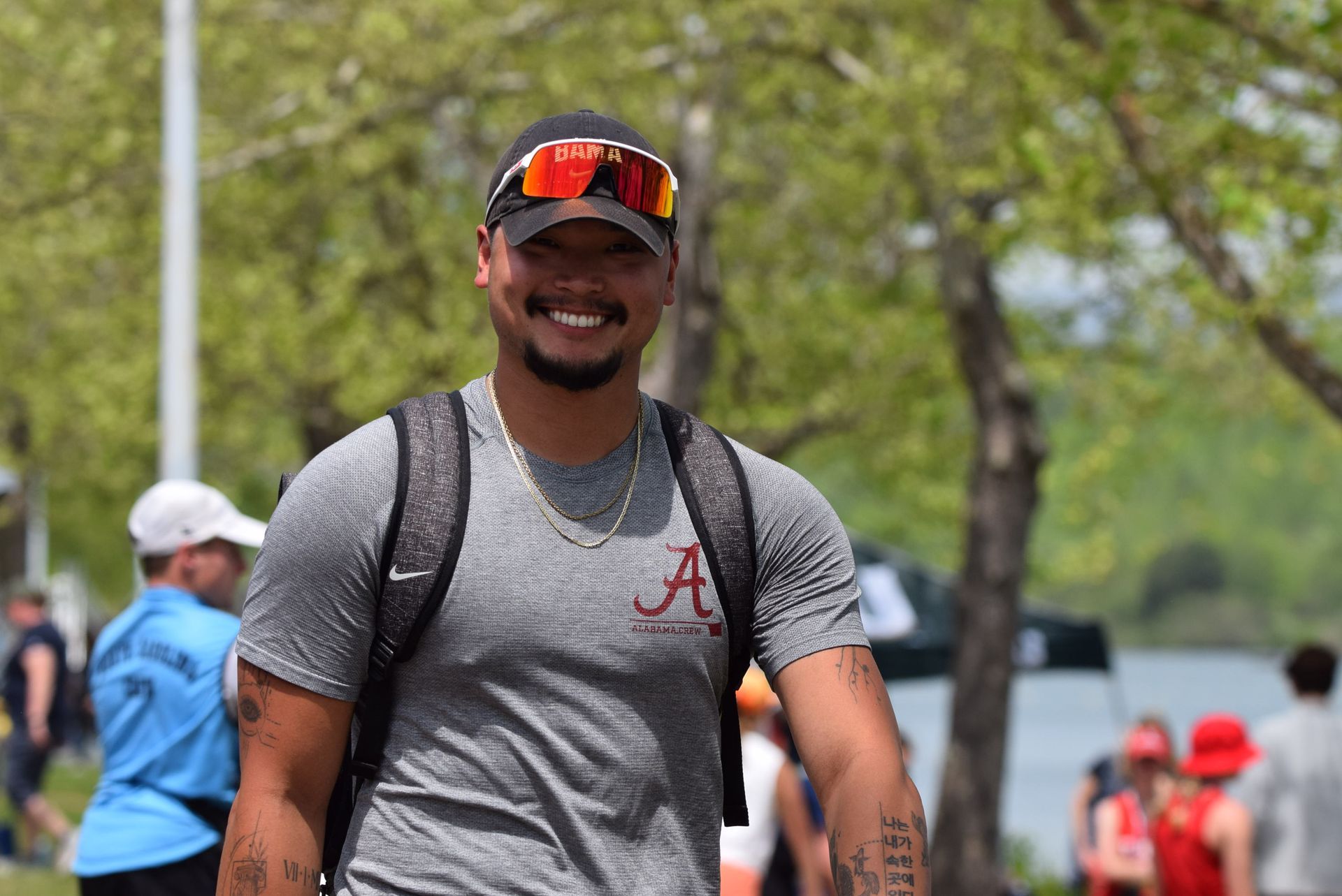 A smiling man wearing a gray Alabama shirt and sunglasses with a backpack walks outdoors.
