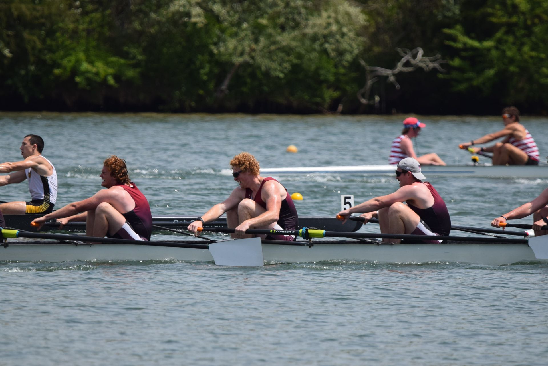 Rowing race on a river. Four rowers in a boat, wearing maroon and white, near other boats in the distance.