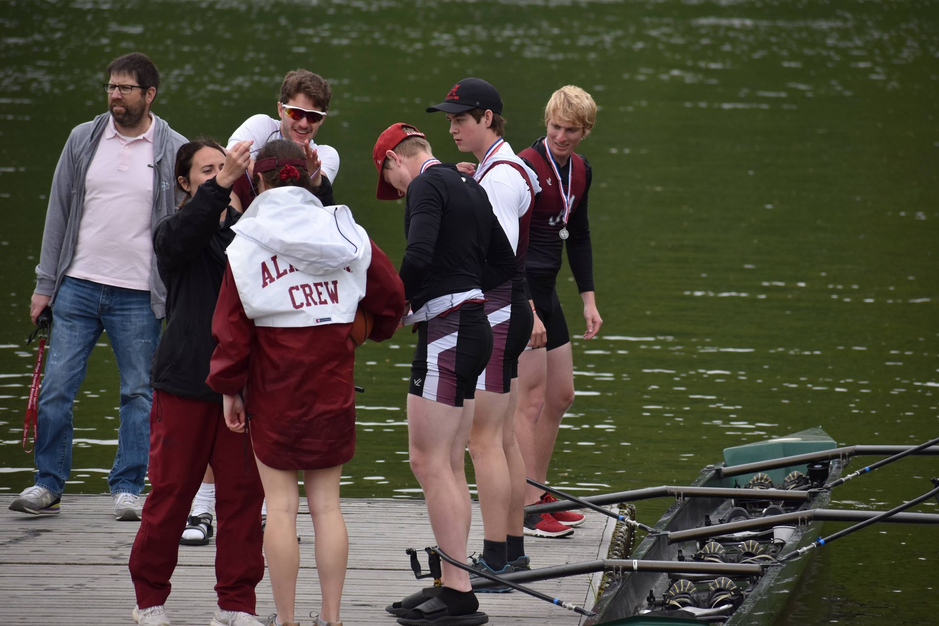 Rowing team members on a dock in maroon and white, with a coach, possibly after a race.