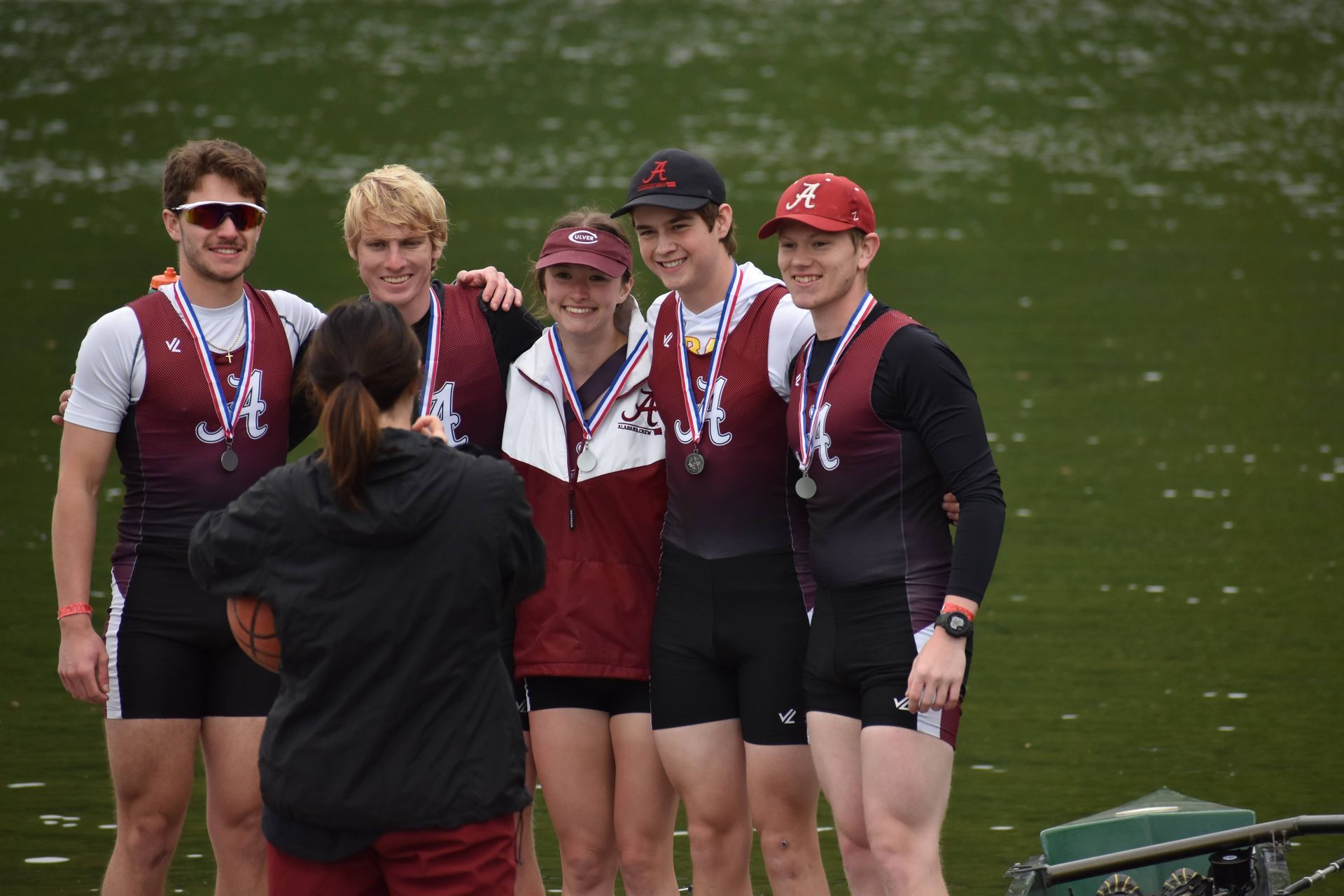Rowing team posing for a photo outdoors, wearing maroon and white uniforms and medals. Person in foreground takes photo.