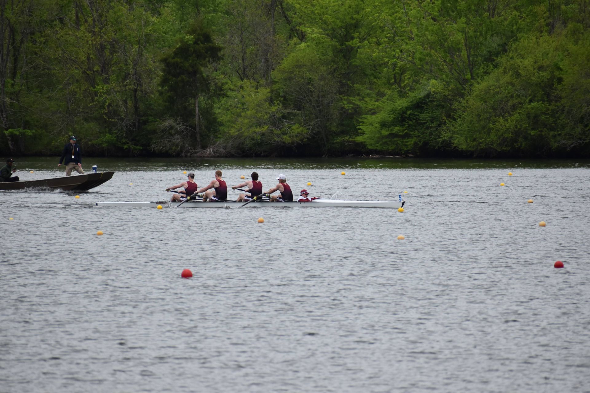 Rowing crew on a lake, four rowers in a boat with a motorboat following; trees in the background.