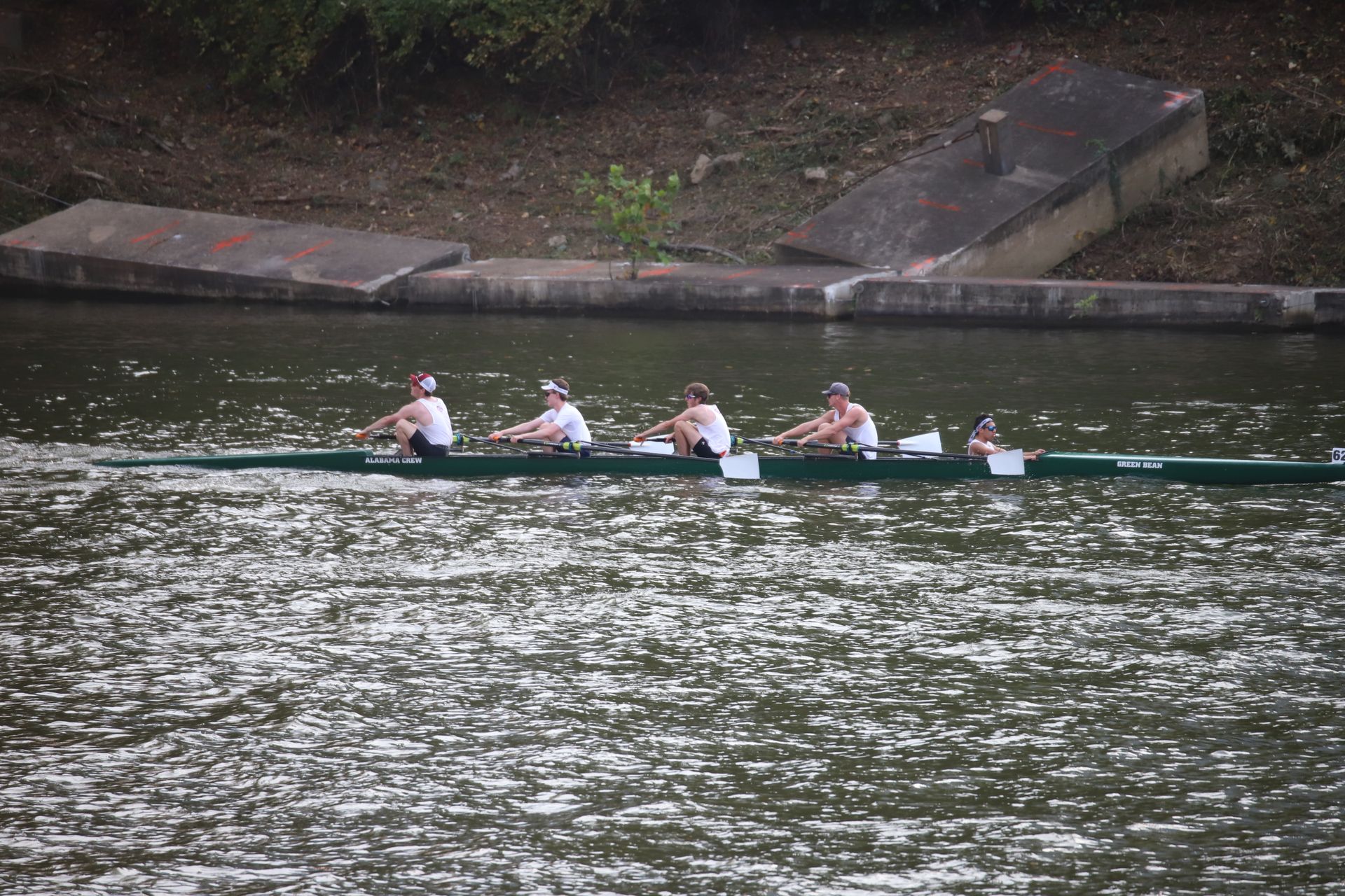 A crew team of four rows on a river. The boat is green, and the rowers wear white shirts and hats. 