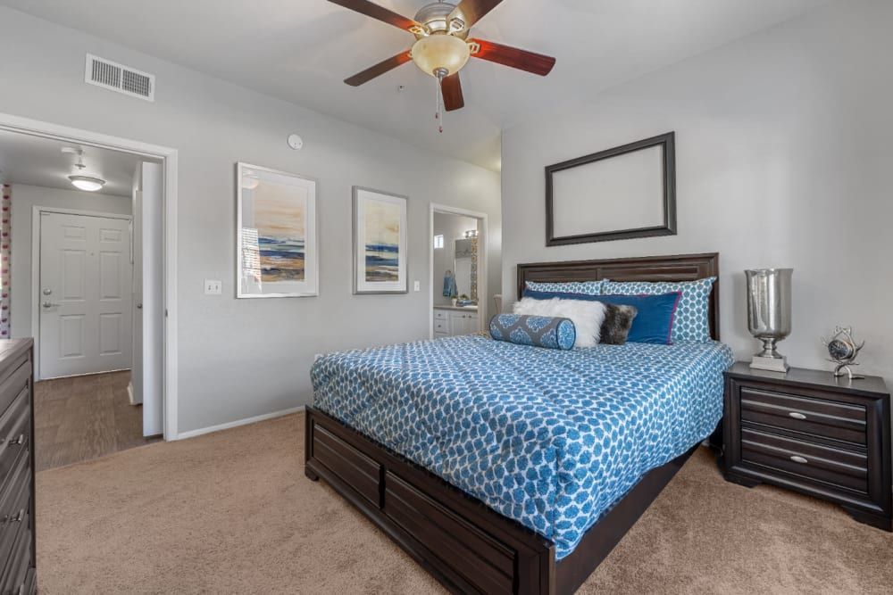A spacious bedroom featuring a large bed with a blue patterned bedspread and multiple pillows, flanked by dark wooden nightstands. A ceiling fan is above, and on the wall opposite the bed is a framed piece of art. The room includes a beige carpet, with a hallway entrance visible to the left leading to another room with a colorful curtain. There's also a mirror and decorative items reflecting a contemporary interior design at Alante at the Islands in Chandler, AZ.