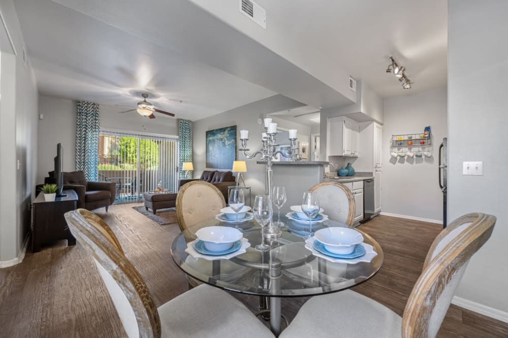 Interior view of a modern apartment living room and dining area. The living room features a brown sofa with matching armchair, a beige accent chair, and a flat screen TV. Natural light streams in from the sliding glass door leading to a balcony with a view of trees. The dining area includes a round glass table set with blue dishes and silverware, complemented by contemporary upholstered chairs. In the background, a white kitchen with cabinets and a breakfast bar can be seen. The walls are decorated with a mix of abstract art and decorative items, and the space is unified with wood laminate flooring at Alante at the Islands in Chandler, AZ.