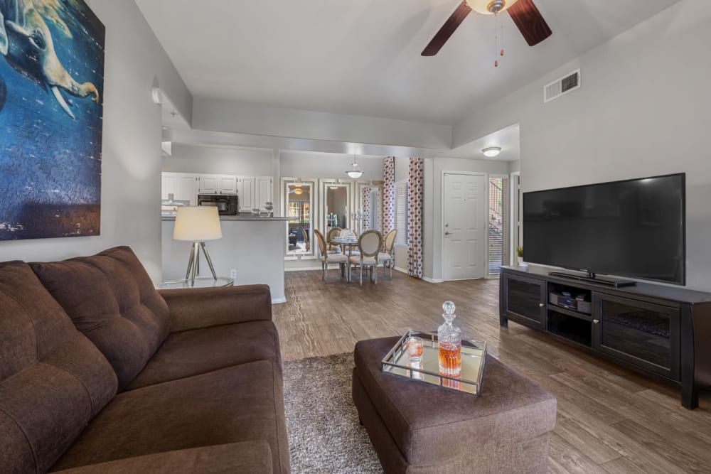 Modern living room with high ceilings and hardwood floors featuring a dark brown sofa with a matching ottoman, a large flat-screen TV on a black media console, and a glass coffee table with decorative items. Artwork hangs on the wall, and the open-concept layout shows a view into the adjoining dining area with a round table and chairs, leading to a kitchen with white cabinetry. Natural light from a window brightens the space at Alante at the Islands in Chandler, AZ.