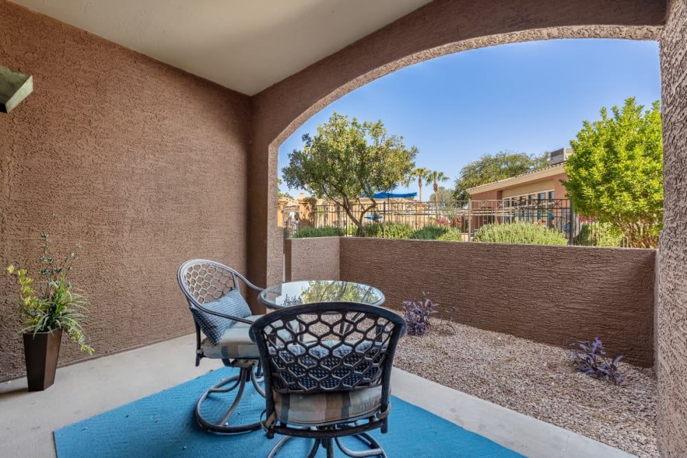 A cozy outdoor patio area under a covered archway, featuring a round table with two wicker chairs. This space offers a view of a neatly landscaped yard with shrubs and trees, as well as a glimpse of the neighboring buildings. The floor is accented with a blue rug, and there is a potted plant to one side, contributing to the tranquil, domestic setting at Alante at the Islands in Chandler, AZ.