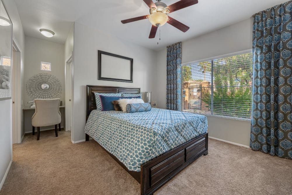 A well-lit bedroom featuring a queen-sized bed with a blue and cream geometric-patterned comforter and several decorative pillows. To the left, there's a small desk with a circular decorative mirror above it and an elegant beige chair. The room has beige carpeting, a ceiling fan with a light fixture, and blue patterned curtains framing a window that looks out to greenery. A framed piece of art hangs above the bed, complementing the room's neutral color palette at Alante at the Islands in Chandler, AZ.