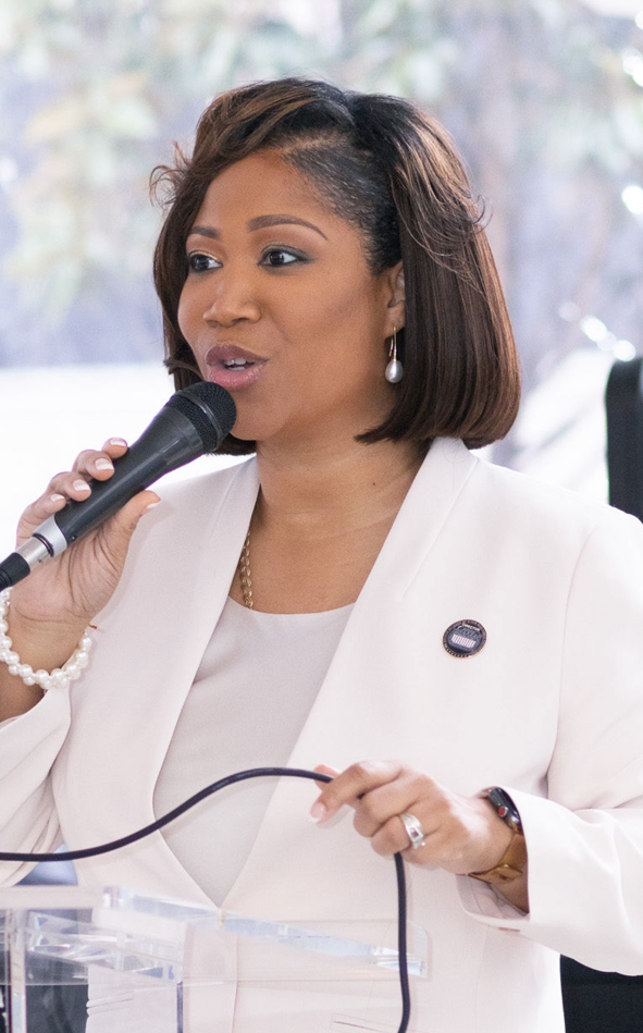 Courtney is speaking into a microphone at a podium, wearing a white blazer and a pearl bracelet.