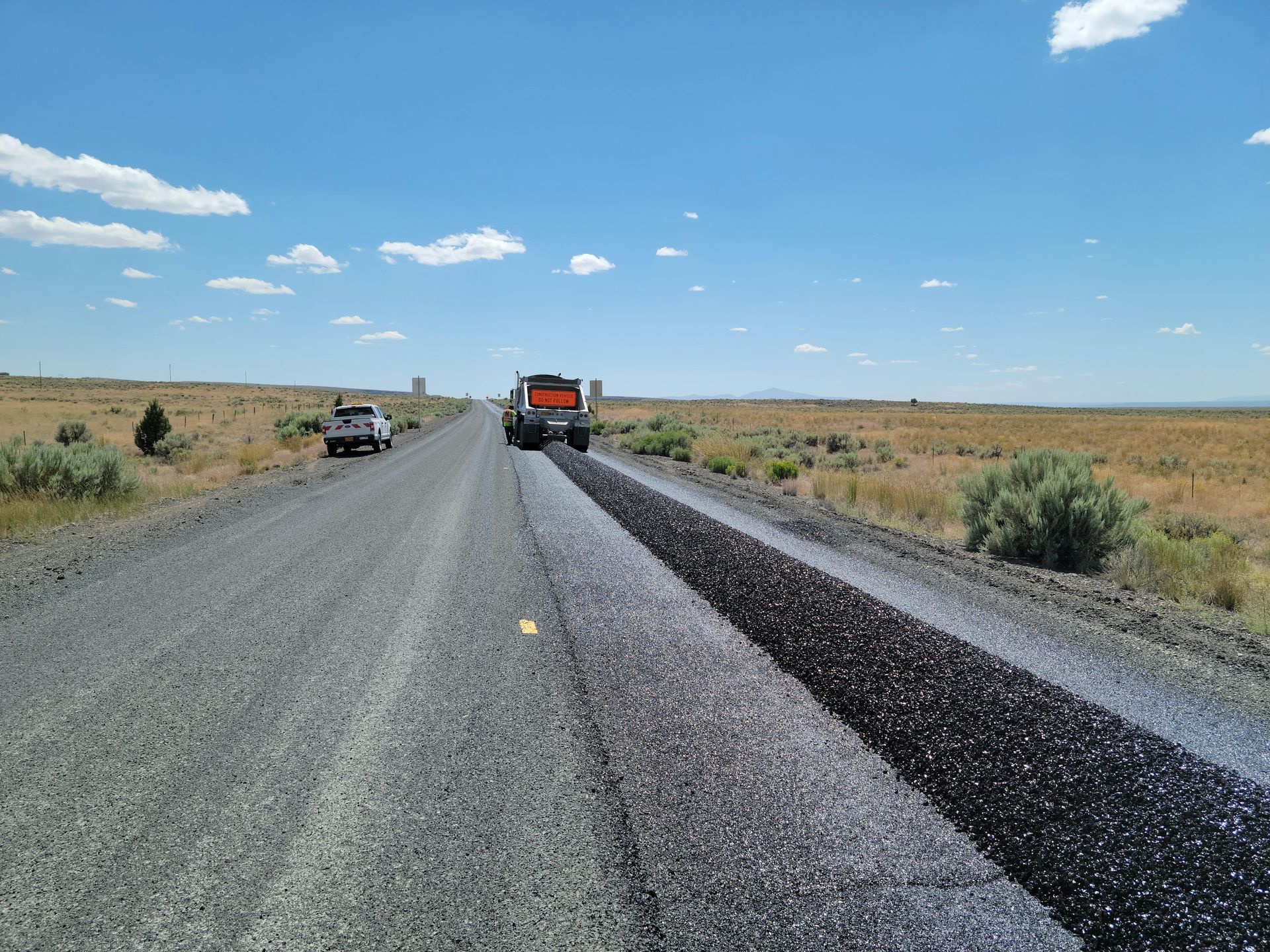 Road paving in progress, a truck applying asphalt on a rural road under blue sky.
