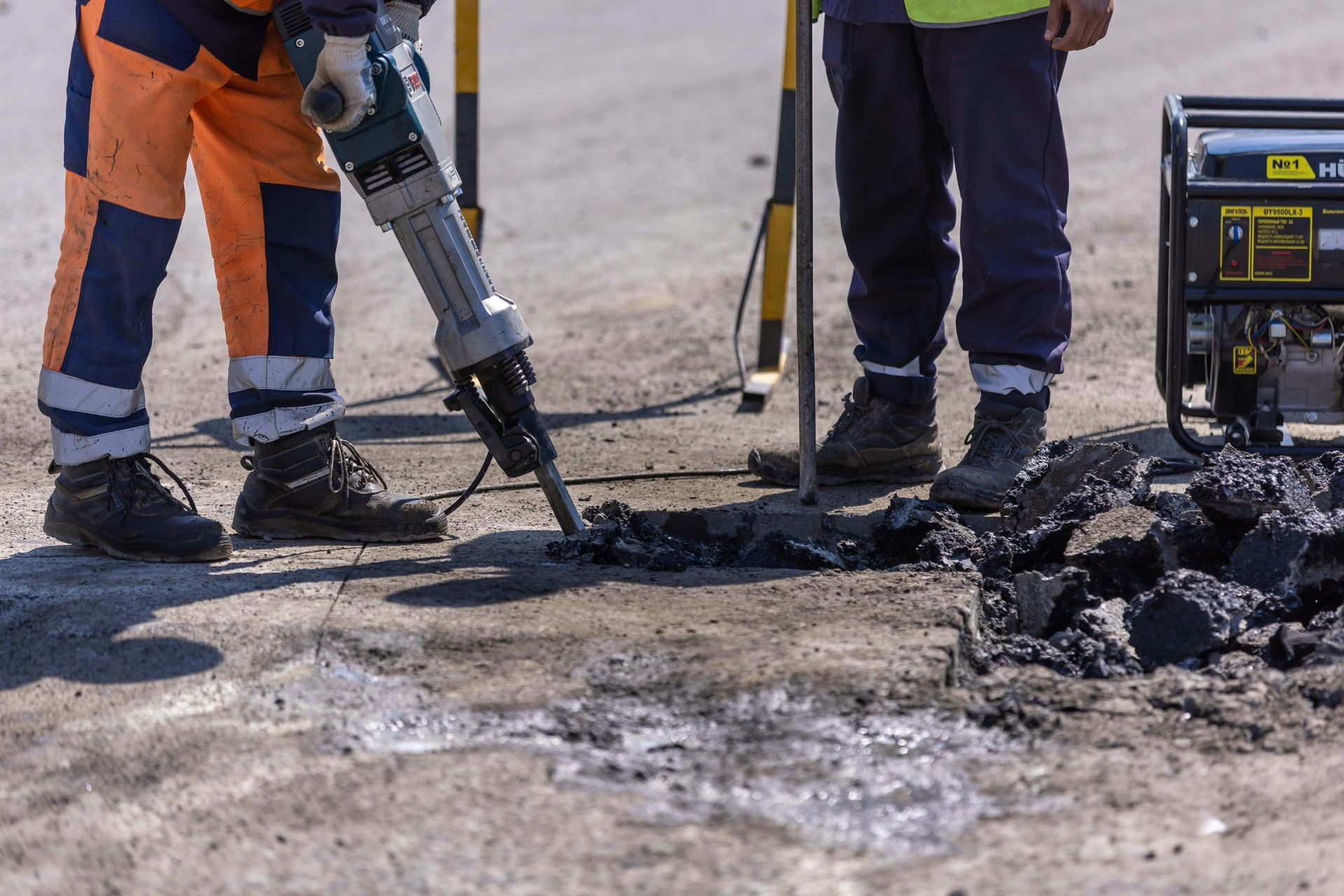 Two workers repairing asphalt with a jackhammer, standing near a generator.