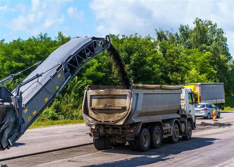 Road milling machine loading asphalt into a dump truck on a sunny day.