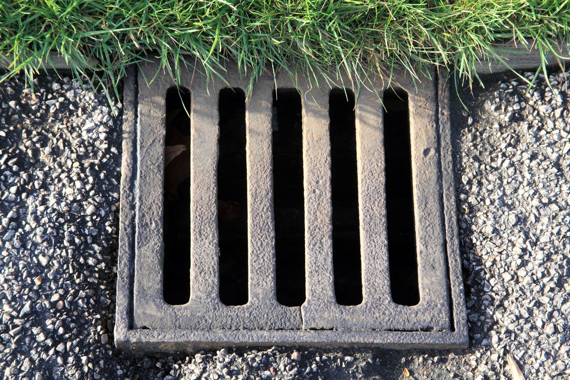 Square metal drain with parallel slots set in asphalt, bordered by grass.