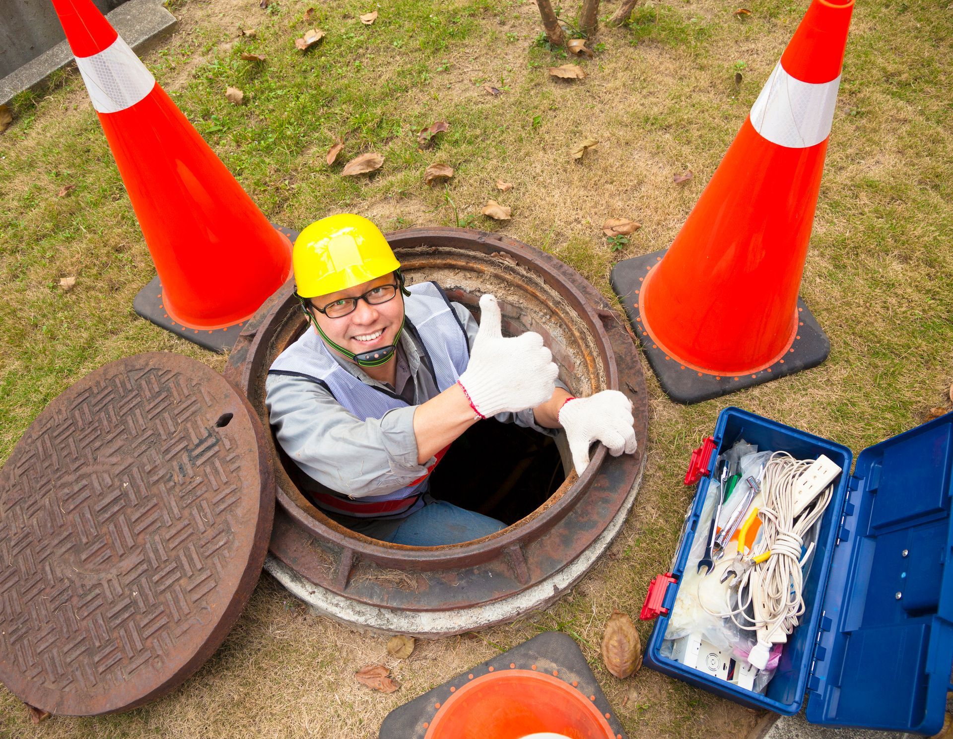 Worker in a sewer, giving a thumbs-up. Orange cones surround an open manhole. Blue toolbox sits beside it.