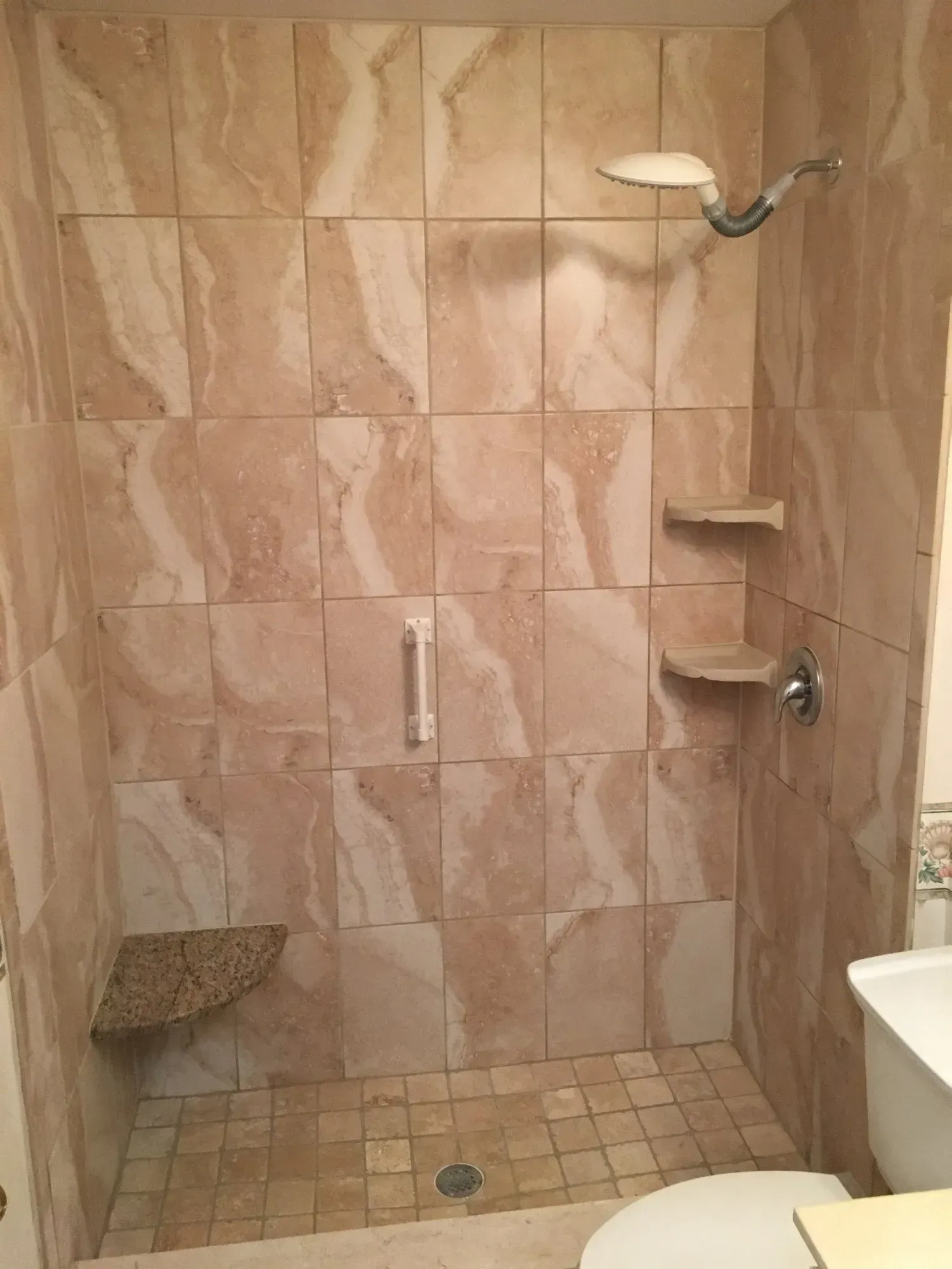 Tiled shower stall with a corner seat, two shelves, and a showerhead. The tiles are beige and the floor has a drain.