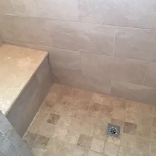 Tiled shower with a built-in bench. Beige stone tiles on the walls and floor, with a drain.