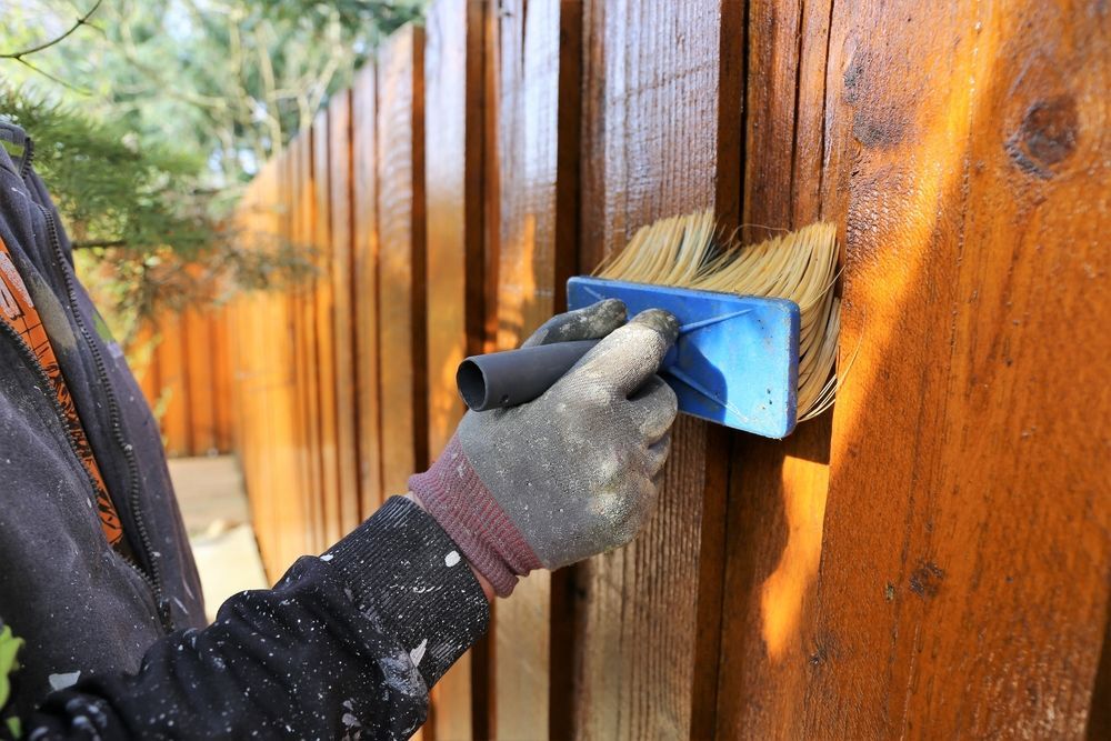 Gloved hand sanding a wooden fence with a blue sanding block outdoors