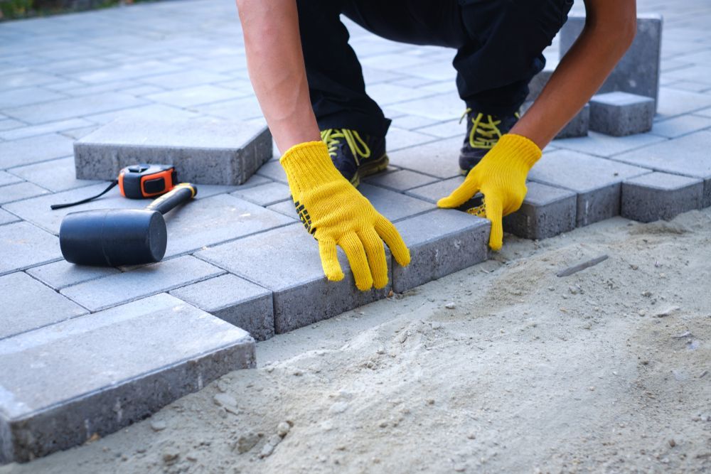 Person in yellow gloves aligning gray paving stones on a walkway with a rubber mallet nearby