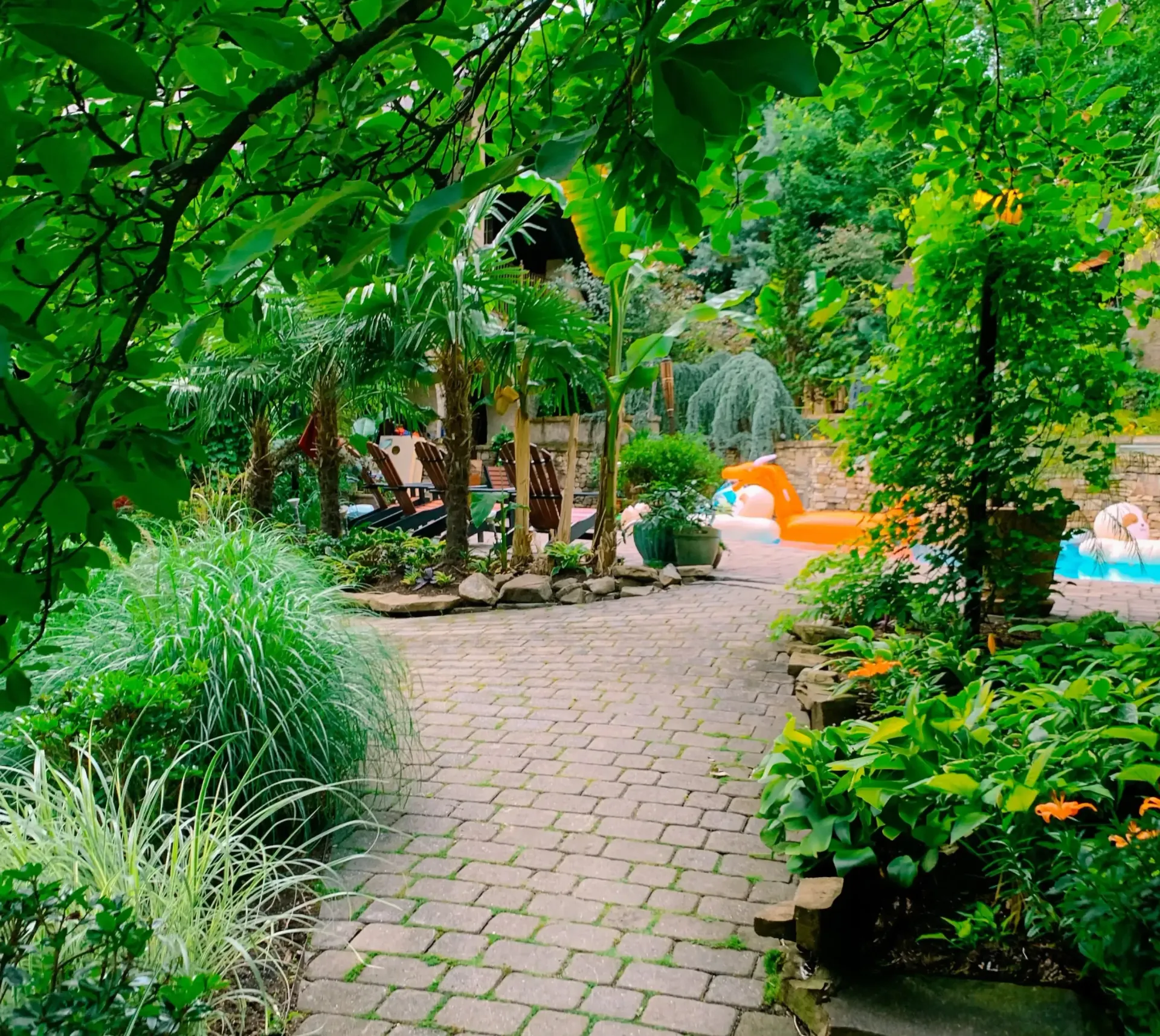A brick walkway leading to a swimming pool surrounded by trees and bushes.