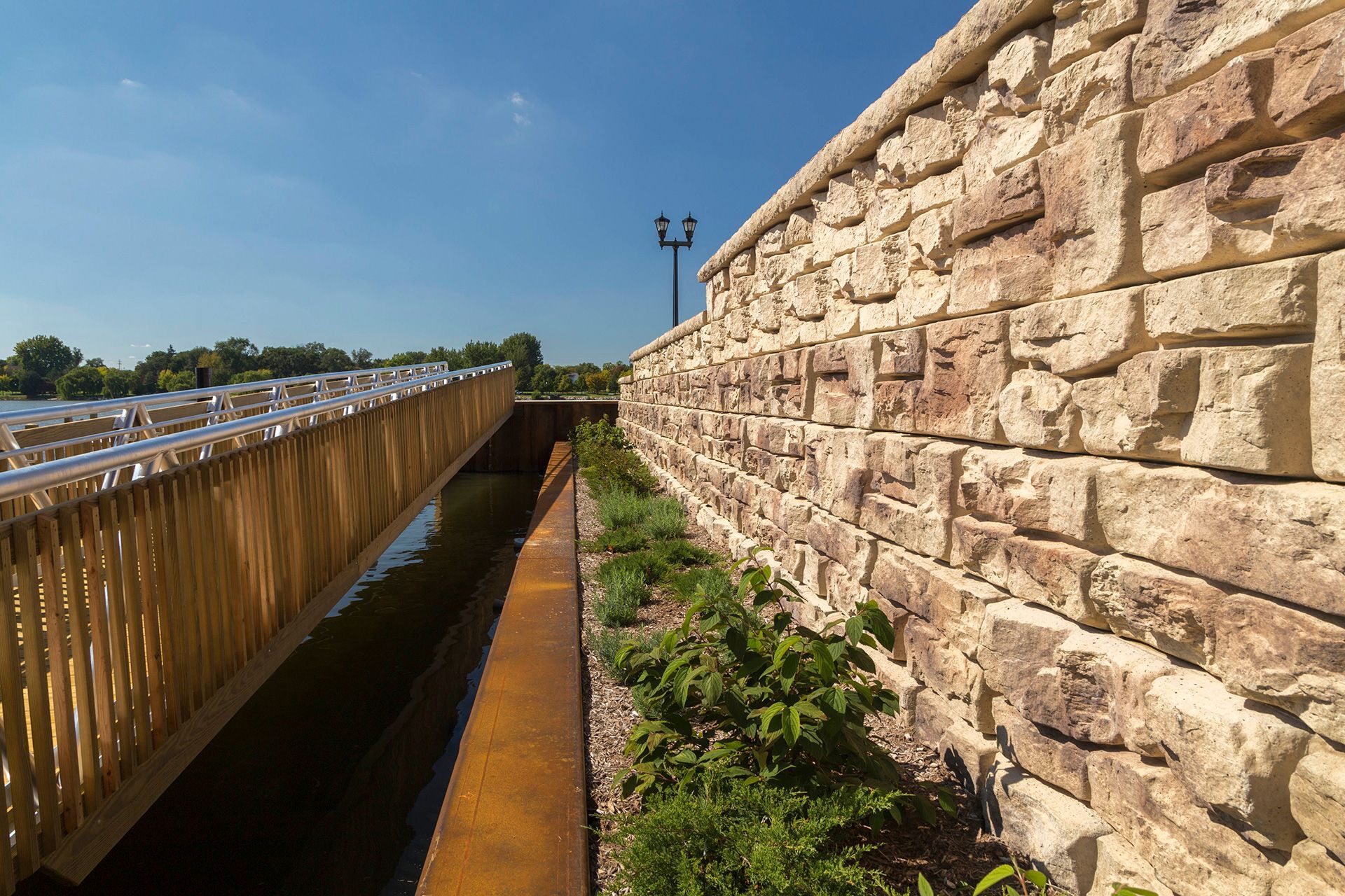 A stone wall along a river with a wooden bridge in the background.