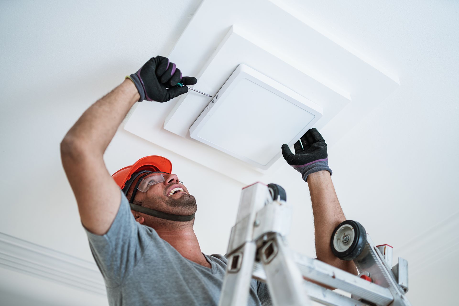 Man on ladder installing ceiling light, wearing gloves and hard hat. White ceiling and light fixture.