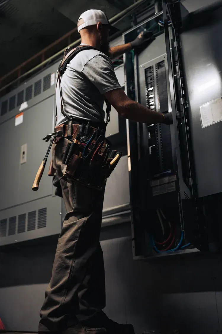 Electrician working on an electrical panel, wearing a tool belt, in a dimly lit industrial setting.