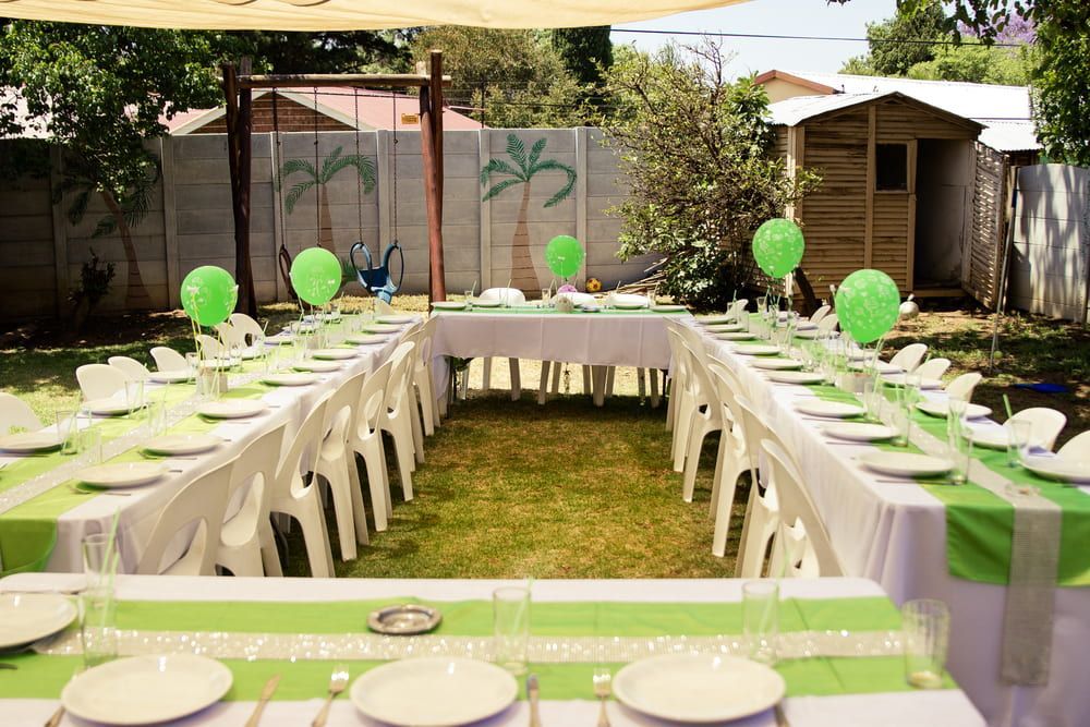 Outdoor party table setup with green balloons, white chairs, and long tables in a backyard garden
