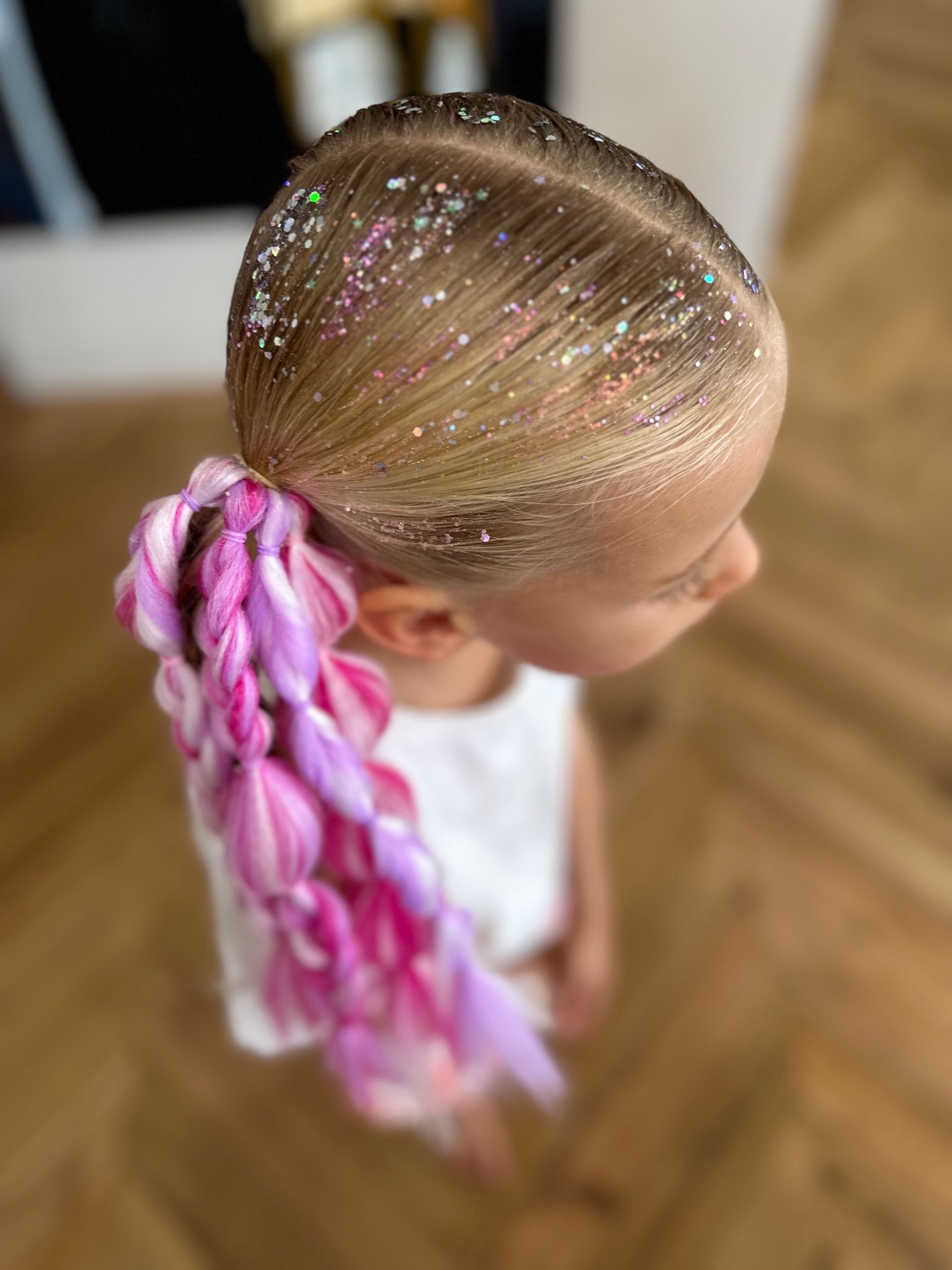 Child with glittery pink braided ponytail viewed from above, standing on a wooden floor. β Sweet Parties & Braids in Tatton, NSW