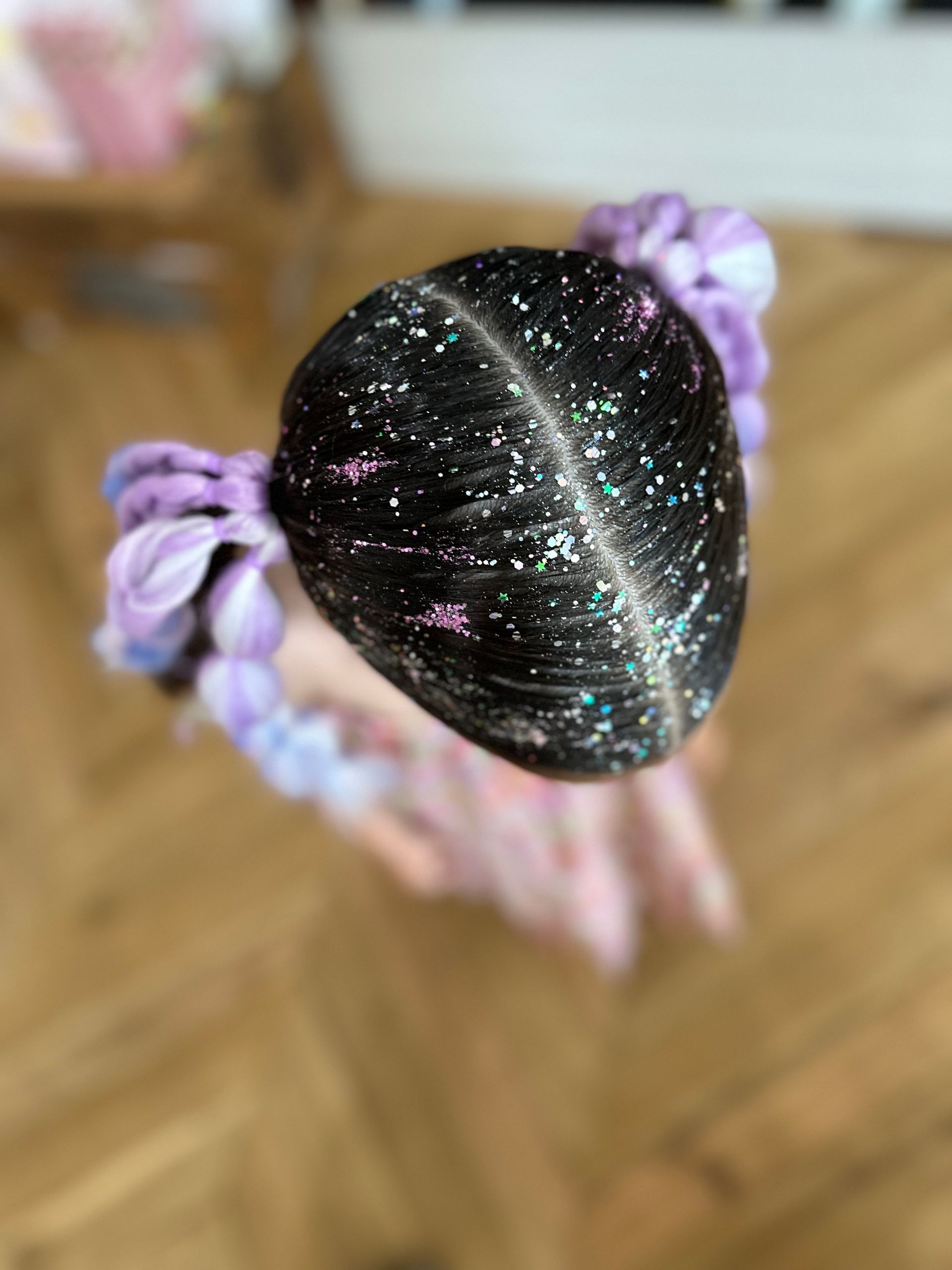 Close-up of a black glittery bun hairstyle with lavender ribbon accents on a blurred wooden floor background β Sweet Parties & Braids in Tatton, NSW