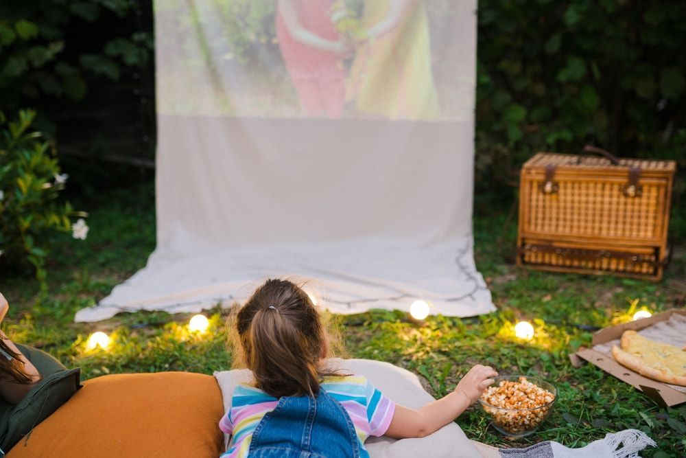 Outdoor movie setup with a projected screen, cushions, string lights, and a wicker basket on grass