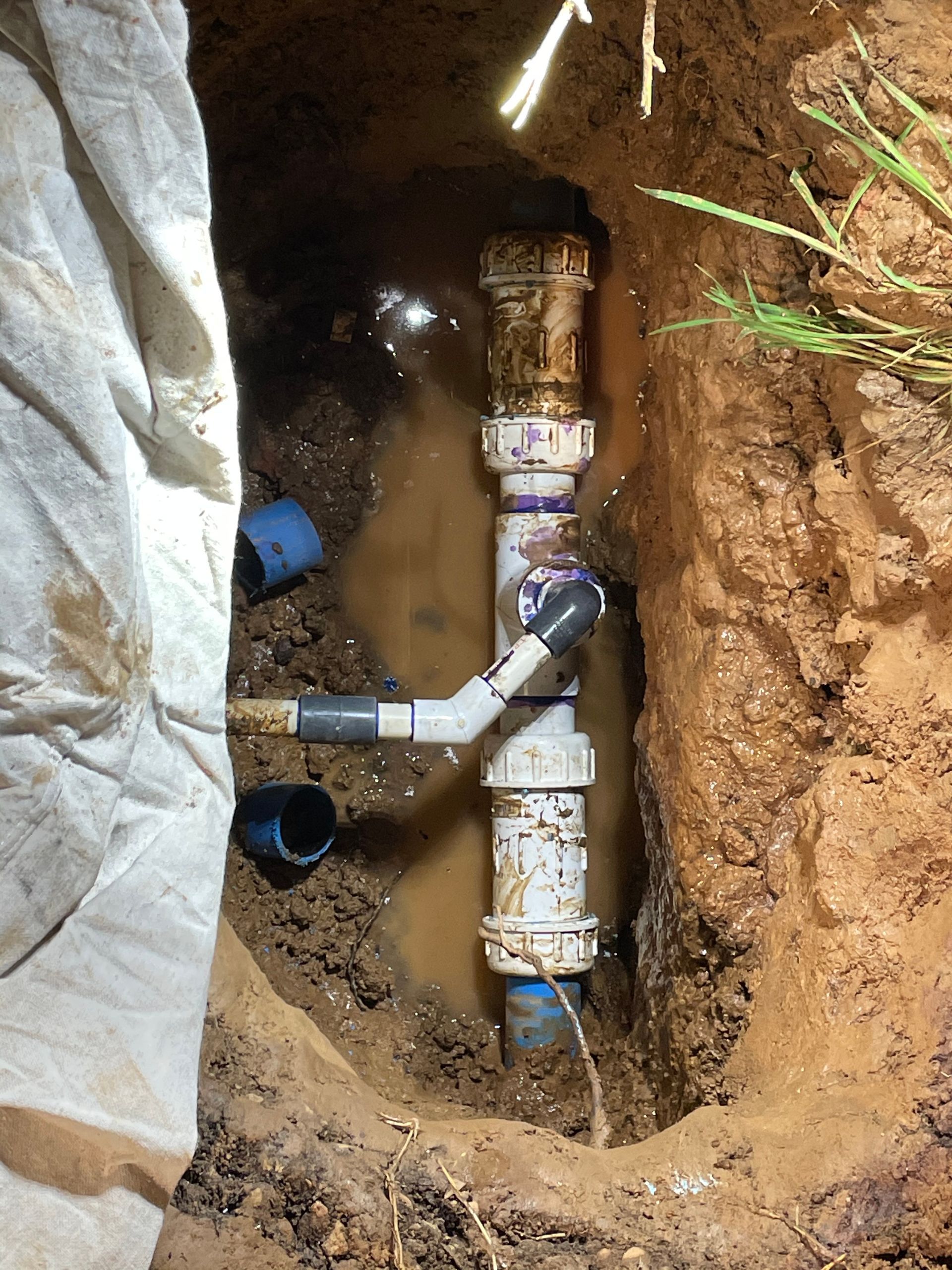 Broken underground PVC pipe in a dirt trench, with a gray tarp and blue fittings nearby.