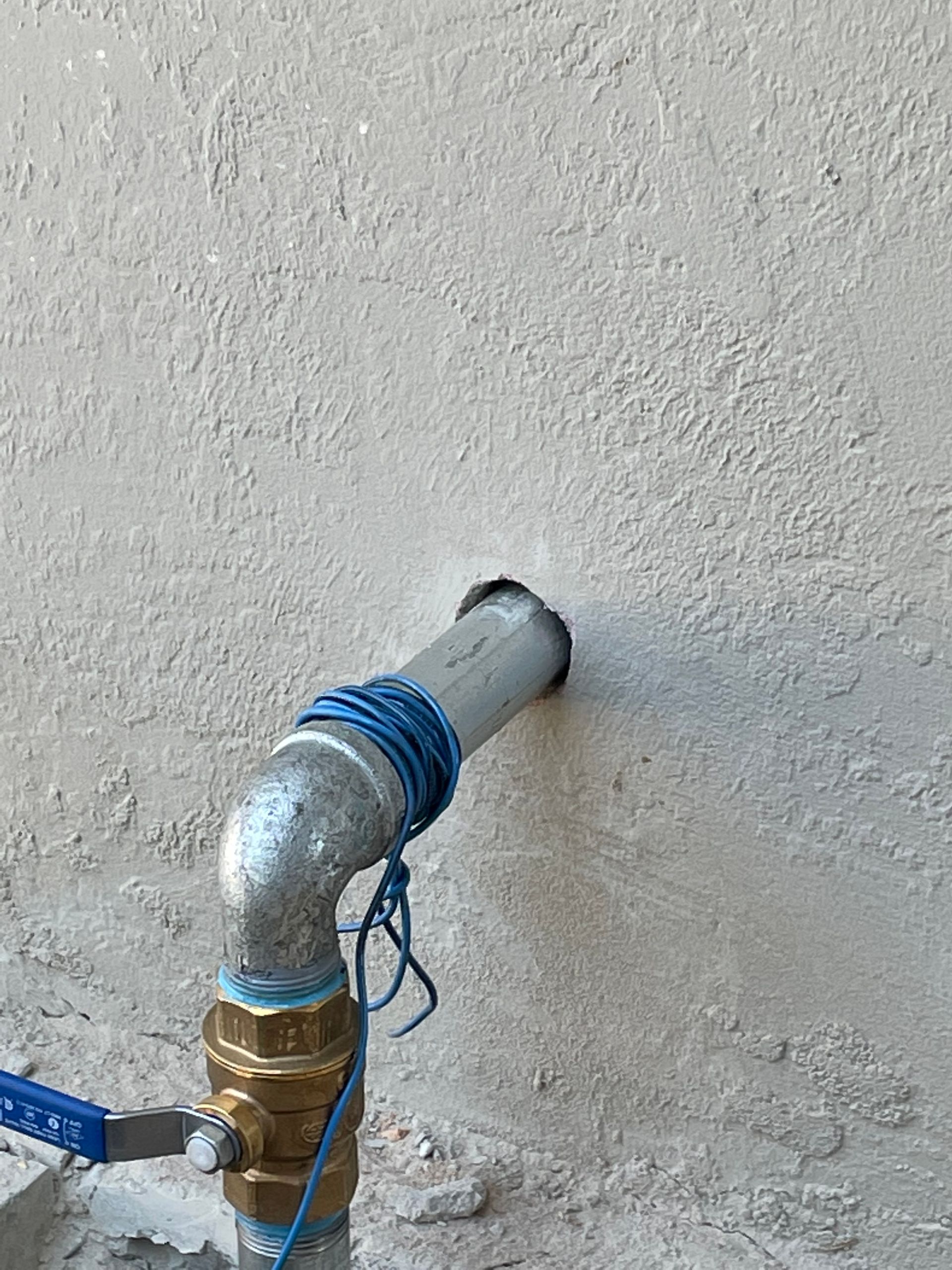 Metal pipe elbow wrapped with blue tape protrudes from a stucco wall.