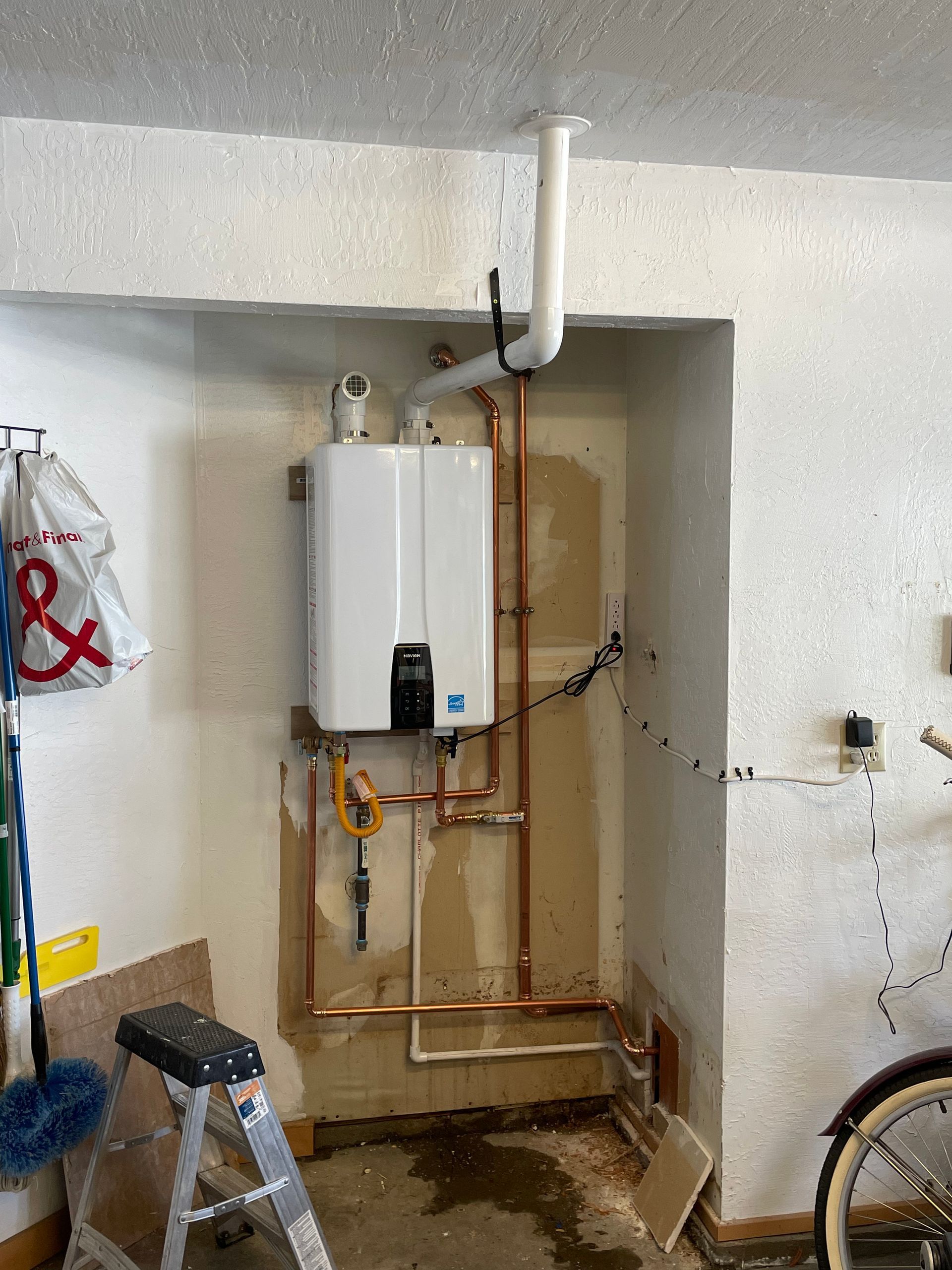 Wall-mounted white water heater with exposed copper pipes in a garage-like utility alcove.