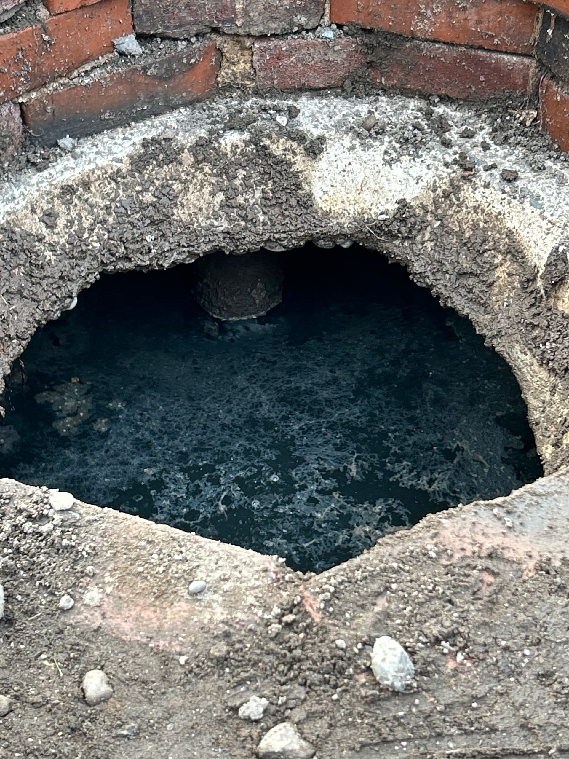 Open circular brick-lined hole with dark water or sludge inside, surrounded by rough concrete and debris.