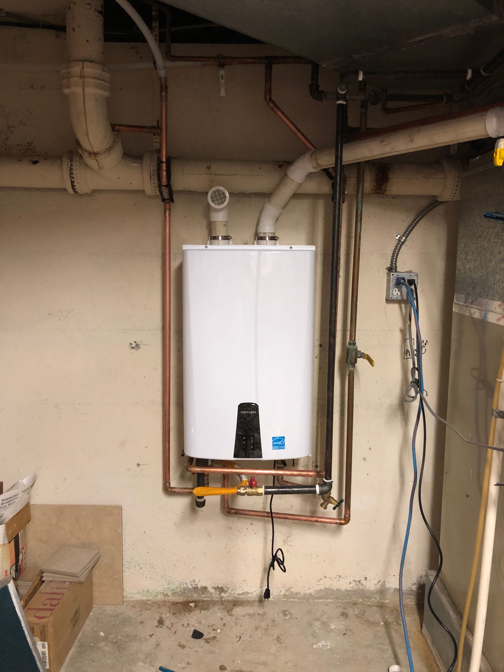 Wall-mounted white water heater with exposed pipes in a utility room, beige walls and concrete floor.