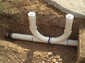 Plumbing pipes installed in a dirt trench, with a white PVC Y-connection and two vertical pipe ends.