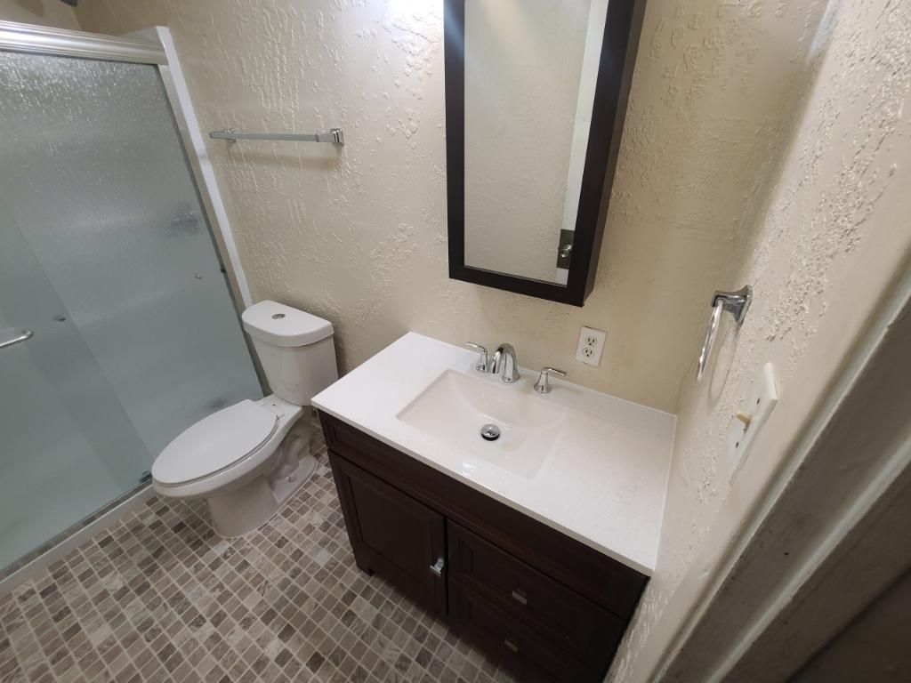 Small bathroom with shower, toilet, and dark vanity with mirror on tiled floor.