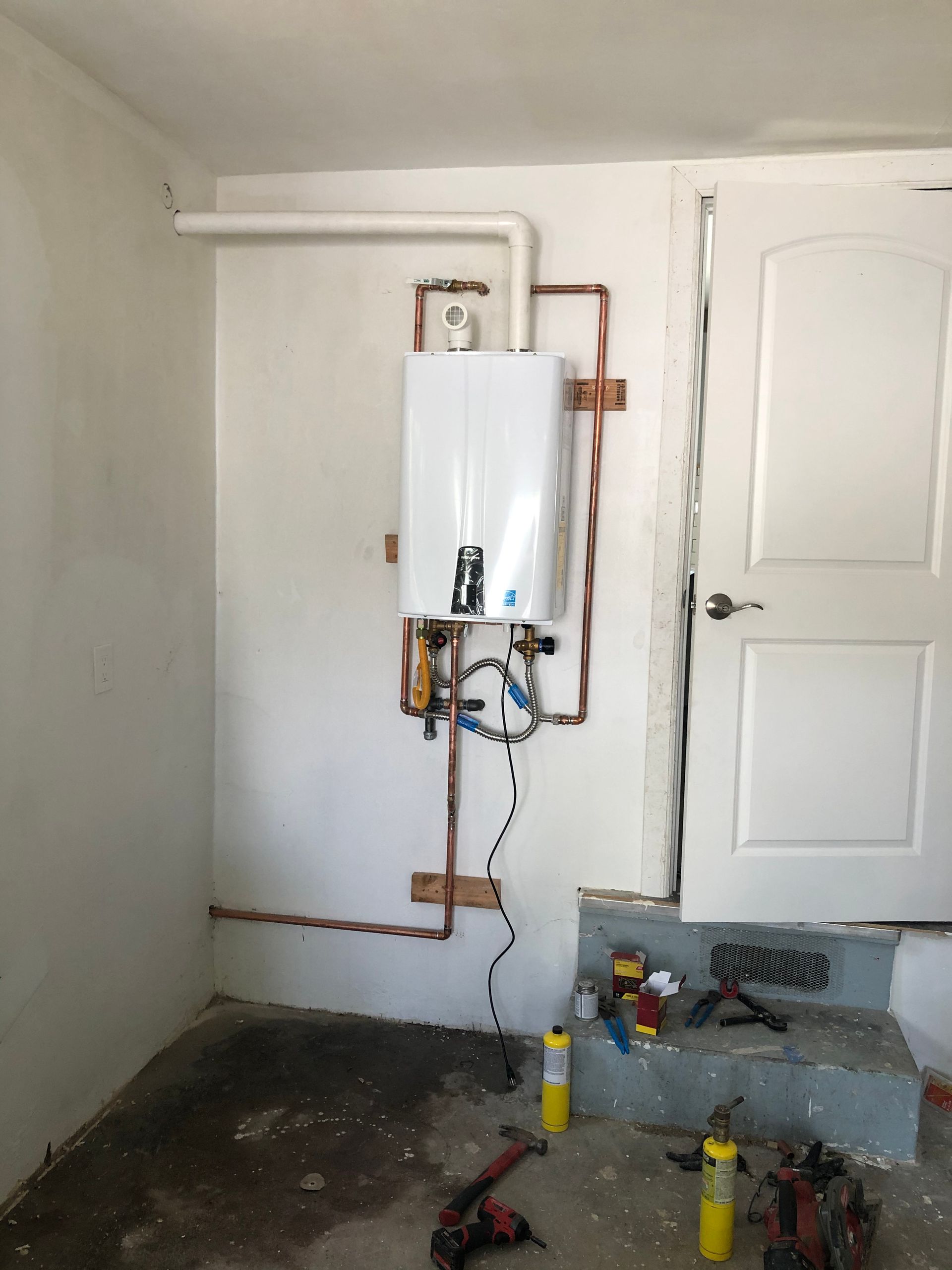 Small white wall-mounted boiler in a plain room with exposed pipes and tools on the floor.