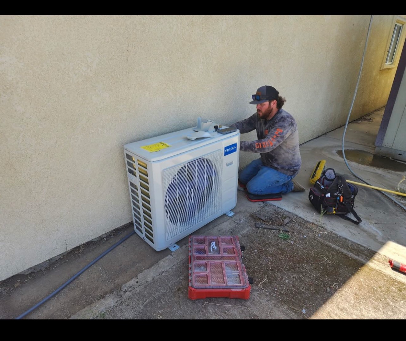 Technician kneeling beside an outdoor air conditioner unit, working with tools against a beige wall.
