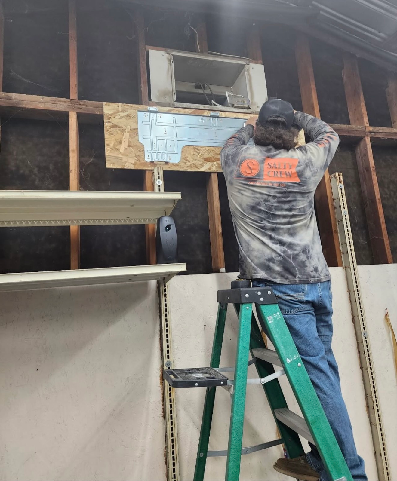 Worker on a green ladder installing a wall-mounted unit in an unfinished room.