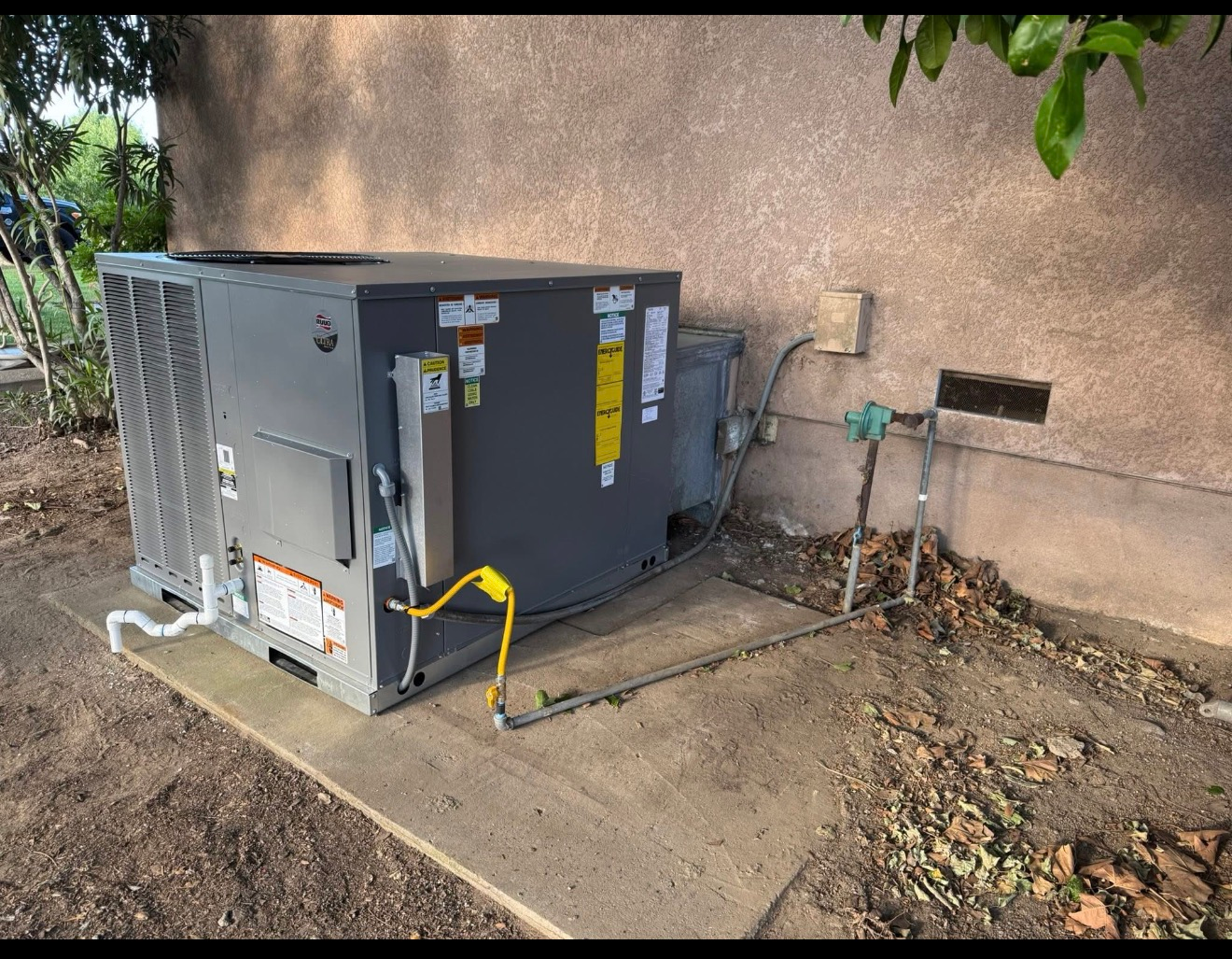 Gray HVAC unit beside a tan wall, with yellow hose and pipes on a concrete pad outdoors