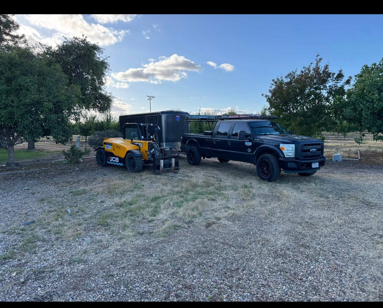 Black pickup truck and yellow tractor parked on gravel with trees and a trailer in the background
