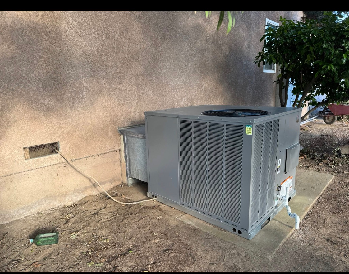 Outdoor HVAC unit beside a beige wall in a dry yard, with a hose and utility lines nearby