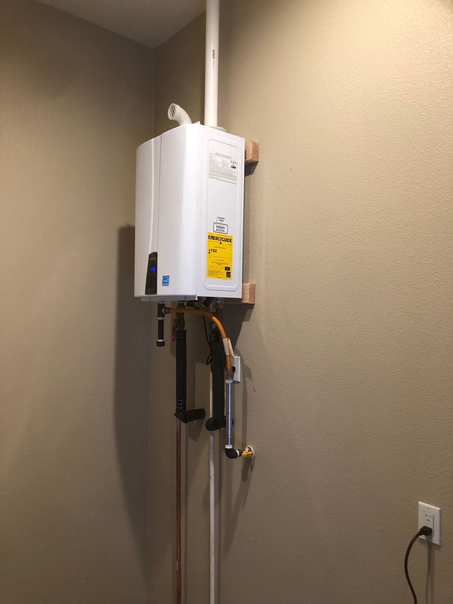 Wall-mounted white water heater with pipes and a yellow warning label in a beige utility room.