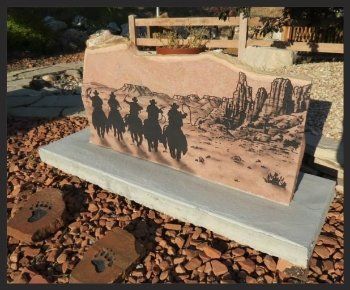 Engraved stone memorial with silhouetted riders, mounted on a concrete base outdoors among rocks.
