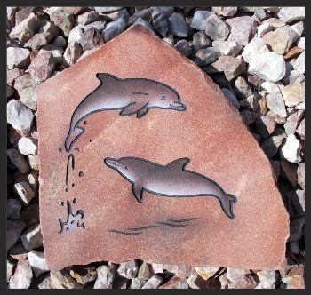 Two dolphin drawings on a pink stone tile among gravel, one leaping above water droplets